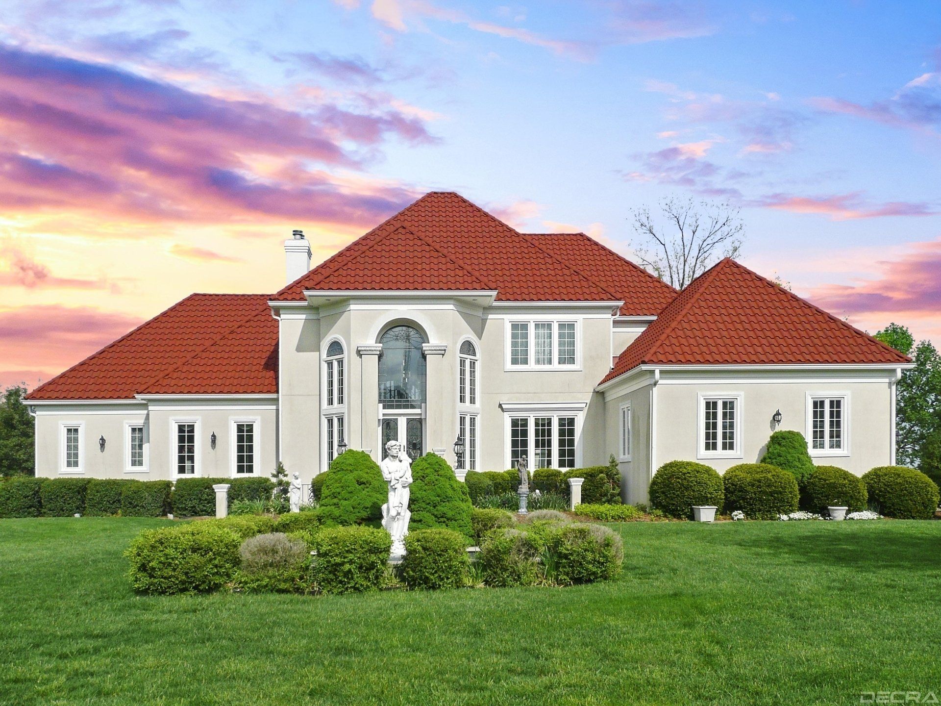 Large cream-colored mansion with red tile roof, lush green lawn, and sculpted bushes under a vibrant sunset sky.