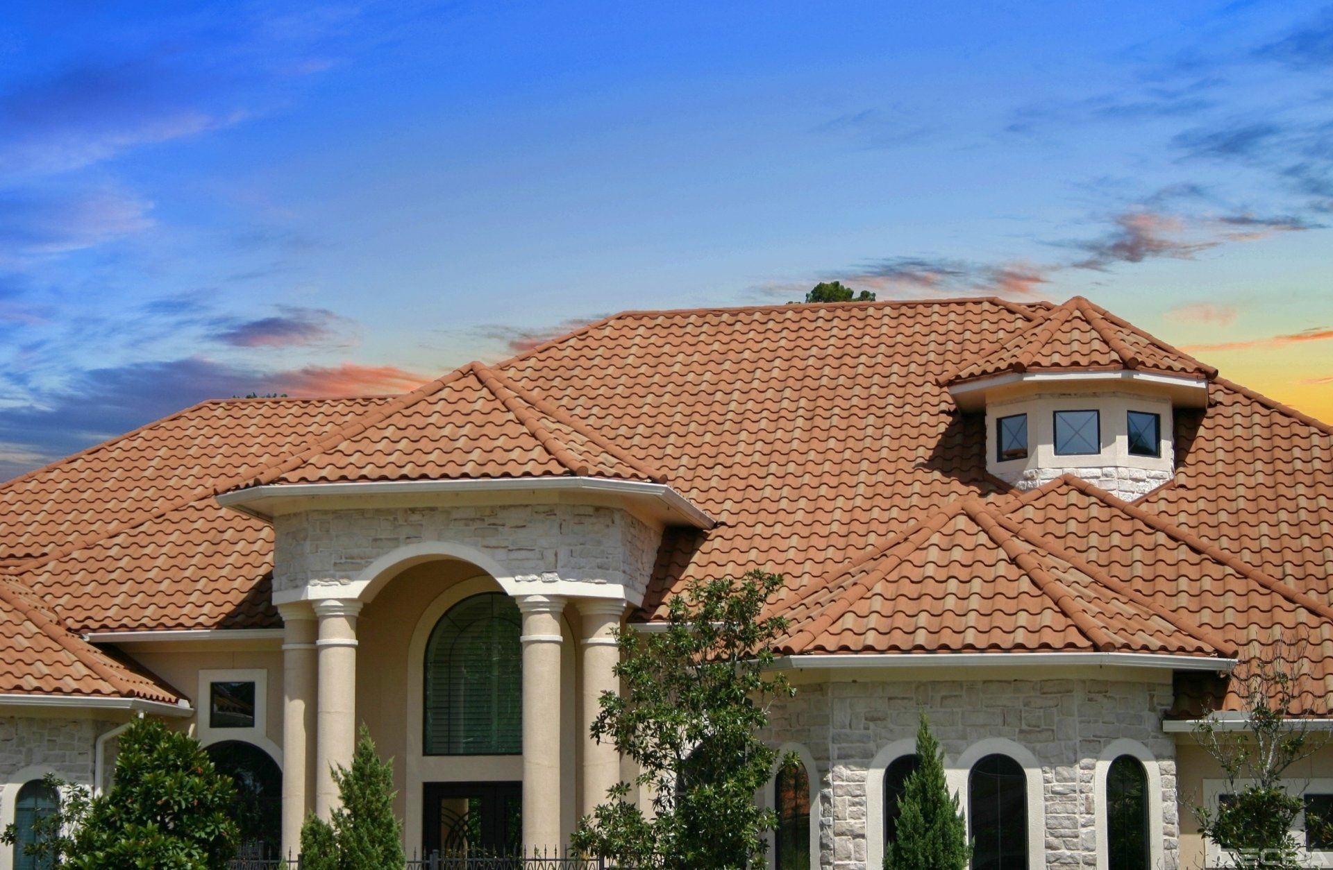 Tan-colored tiled roof of a large house against a blue sky with clouds. White stone facade, arched windows, and columns.