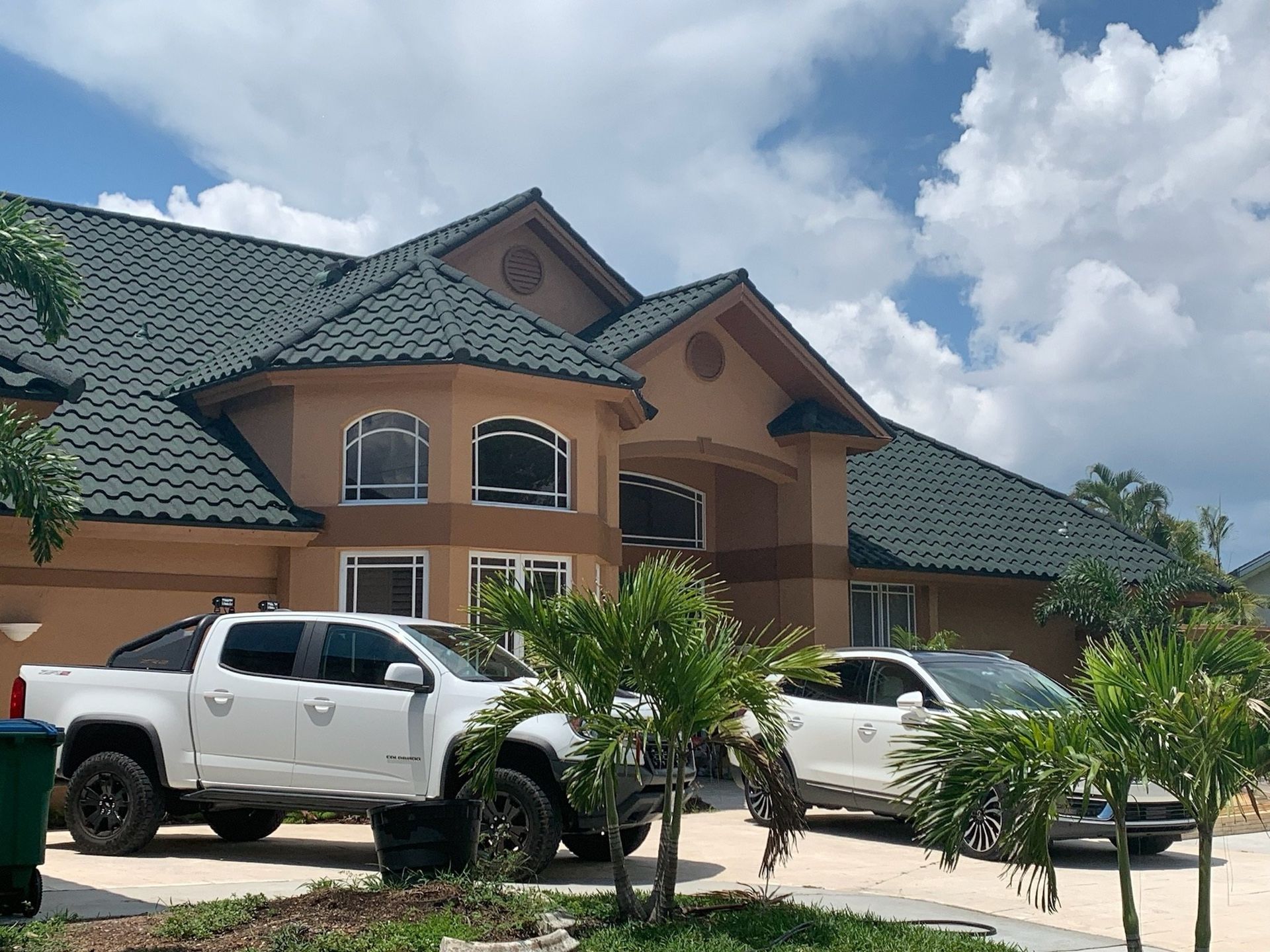 White truck and SUV parked in front of a tan house with a green tile roof on a sunny day.