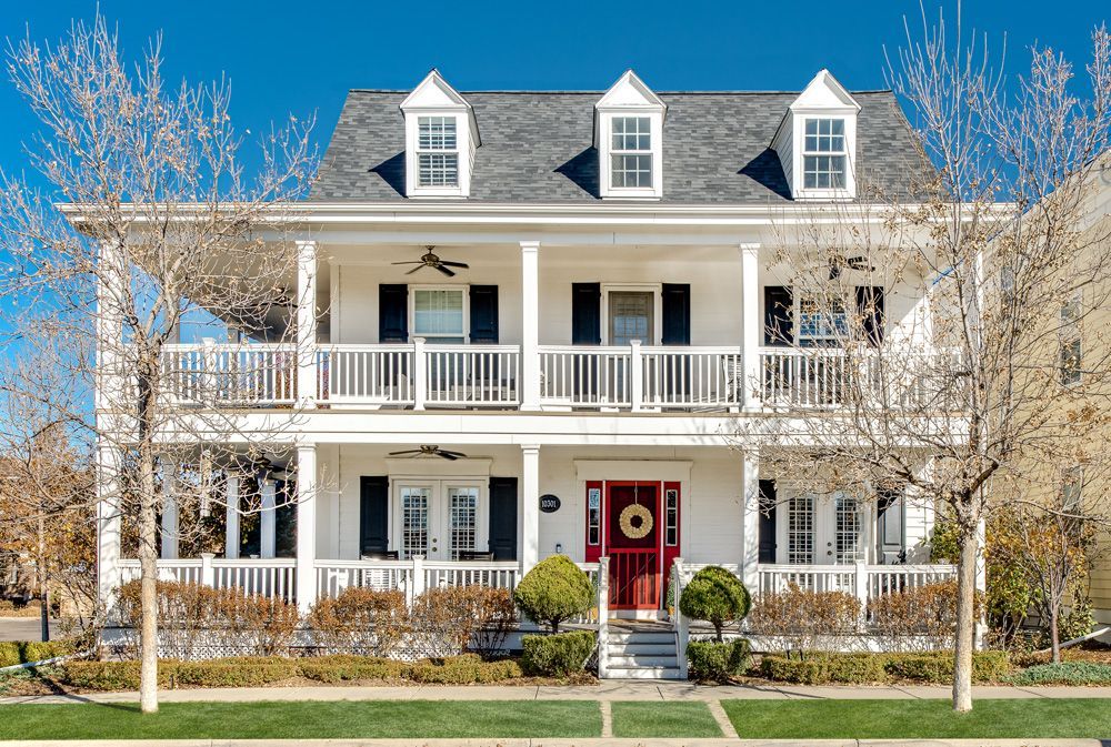 Two-story white house with black shutters, red front door, and a porch. Three dormer windows are on the gray roof.