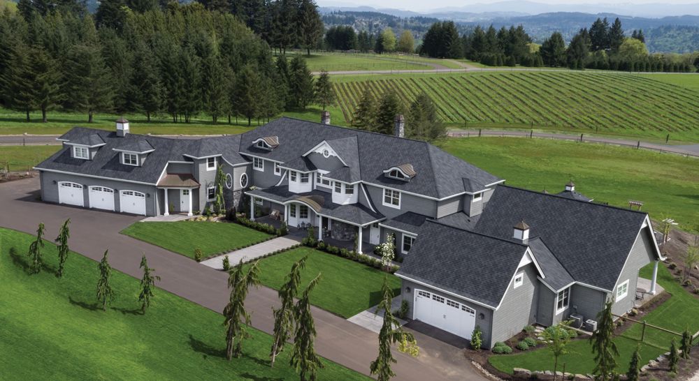Large gray house with multiple garage bays, a long driveway, and a green lawn, set against a backdrop of a vineyard and trees.