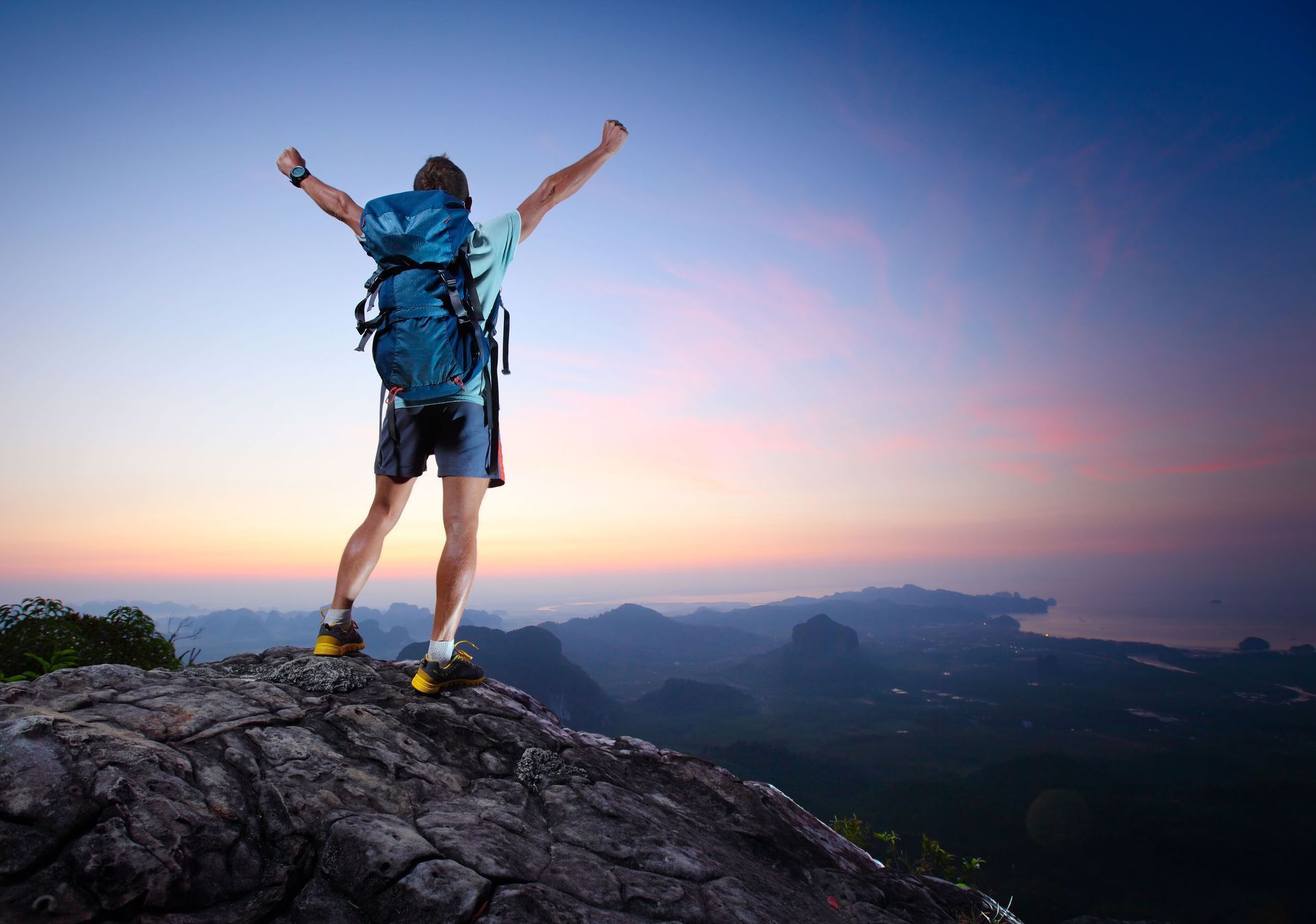 Hiker on mountain peak with arms raised, backpack. Sunset sky, mountain range below.