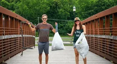 Two people on a bridge holding filled trash bags; cleaning up the environment.