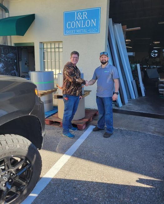 Two men shaking hands outside J&R Conlon, beside a parked truck and store entrance.