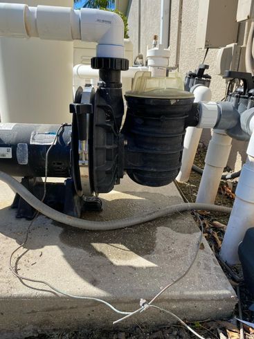 Pool pump system on concrete pad, with pipes, filter, and electrical wiring, in an outdoor setting.