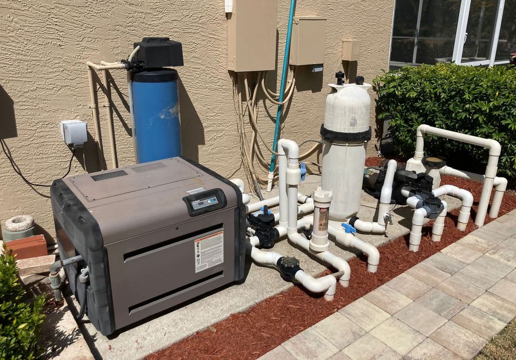 Pool equipment setup: heater, filter, and plumbing against a stucco wall, surrounded by brick and mulch.