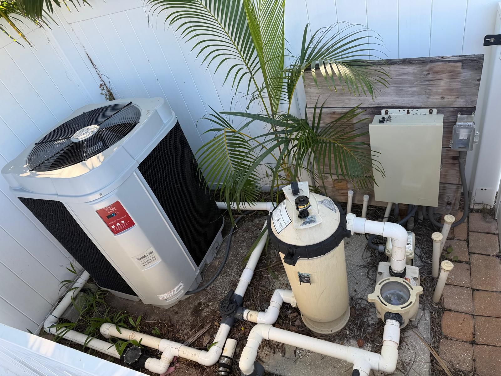 Pool equipment next to a white fence. A heat pump, filter, pump, and electrical box are visible.