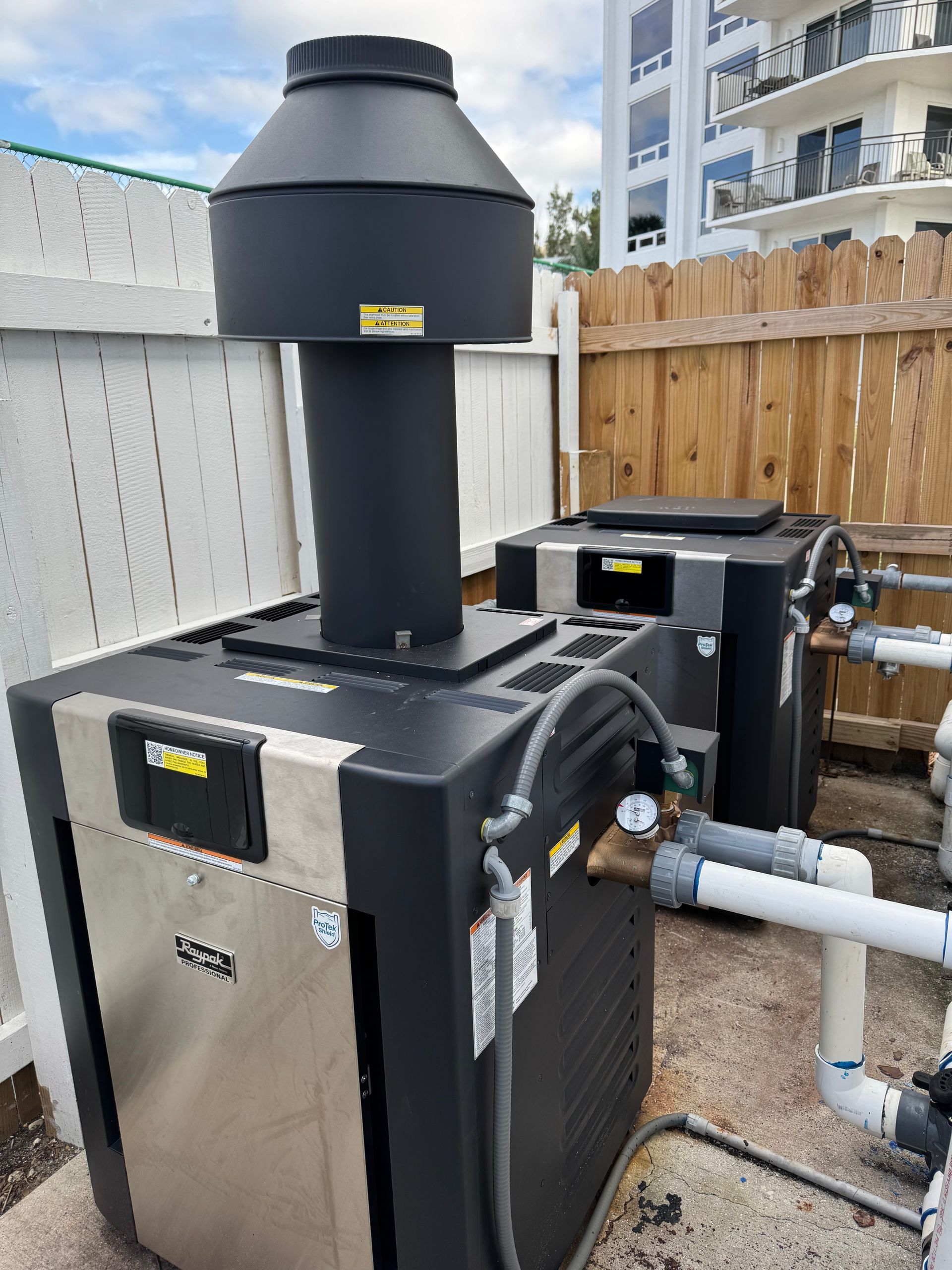 Two pool heaters with black tops and silver sides next to a white fence.