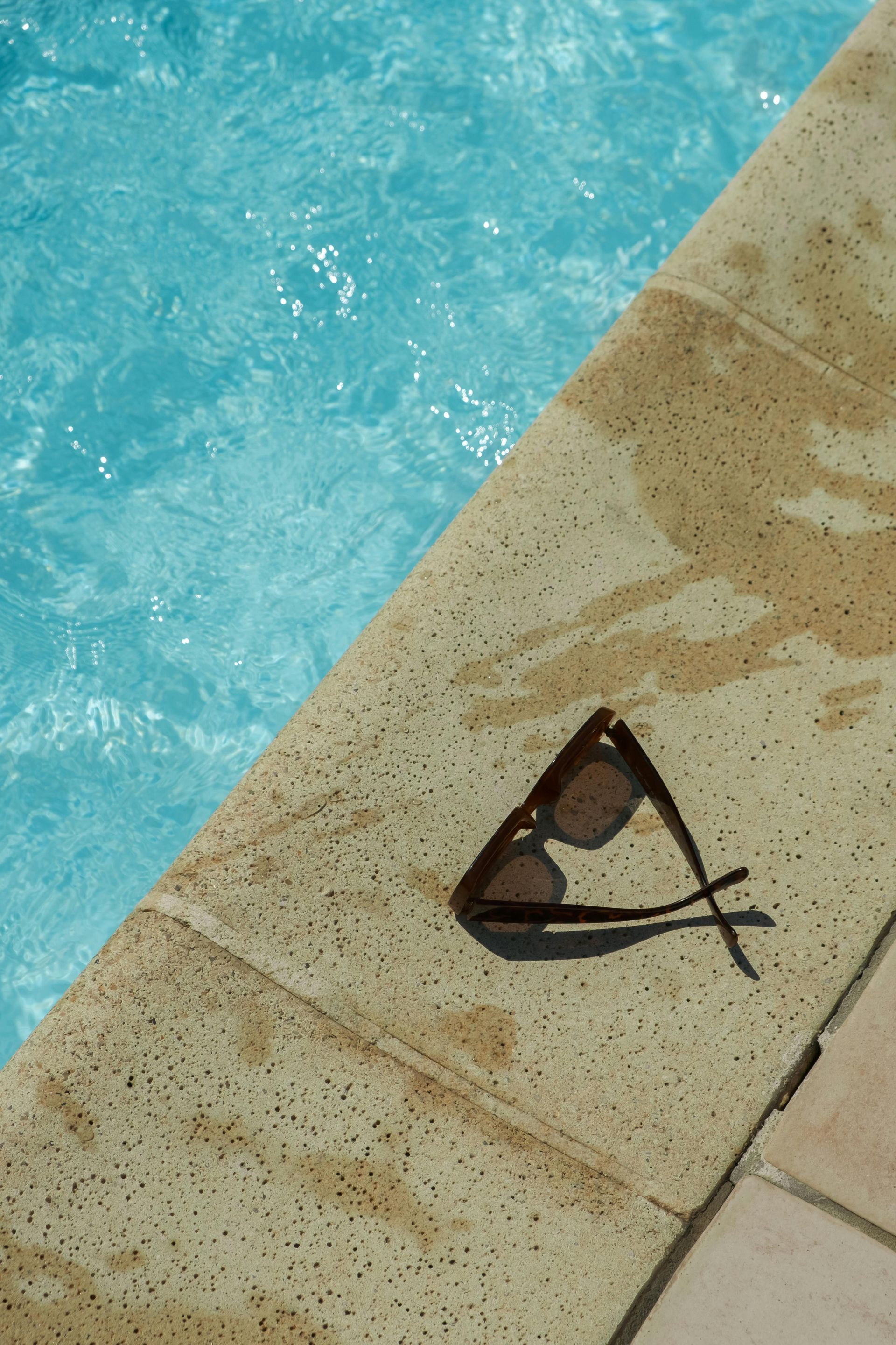Sunglasses resting on the edge of a light-colored pool, with blue water in the background.