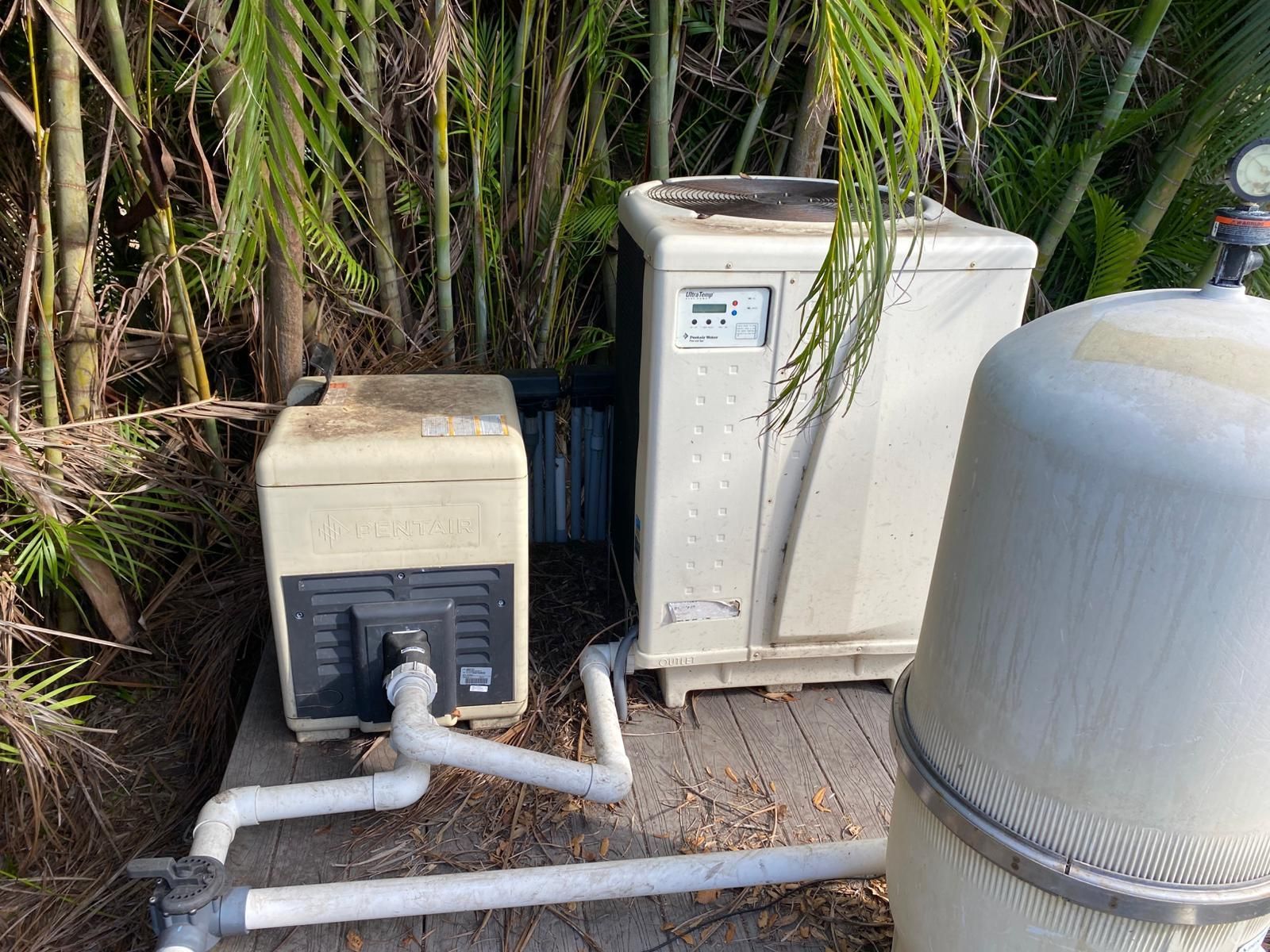 Pool equipment: pump, heater, and filter on a wooden platform surrounded by foliage.
