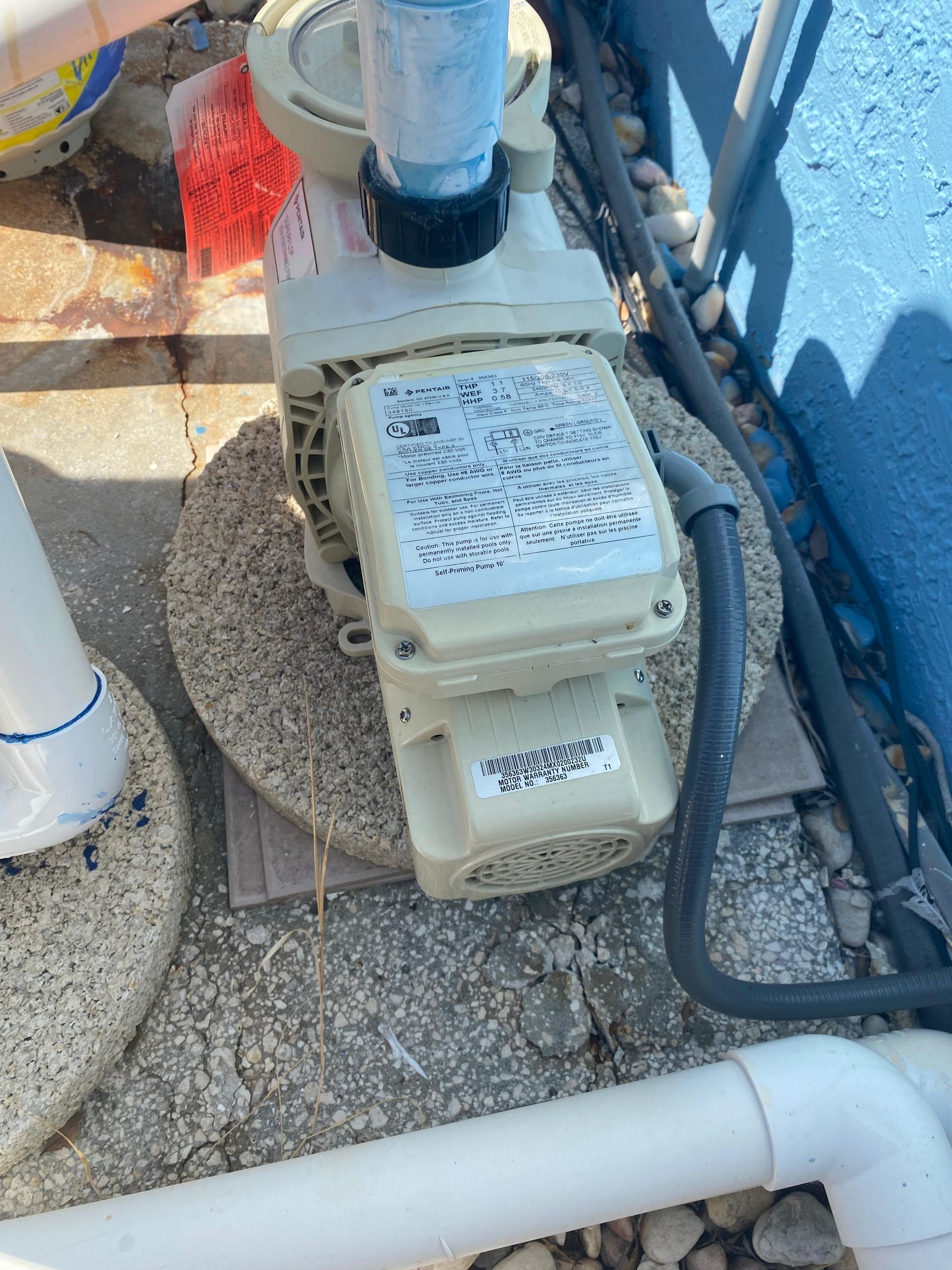 Pool pump on a circular concrete base, connected to white pipes and gray electrical conduit, near a blue wall.