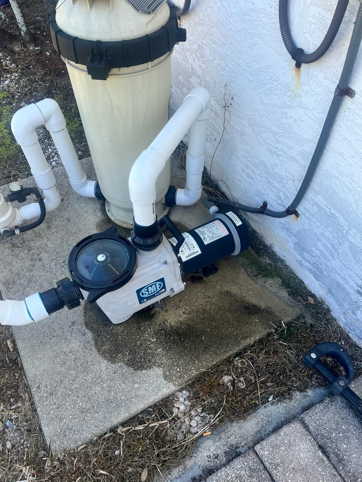Pool pump and filter system near a textured wall. White pipes connect to a tan filter and a gray pump.