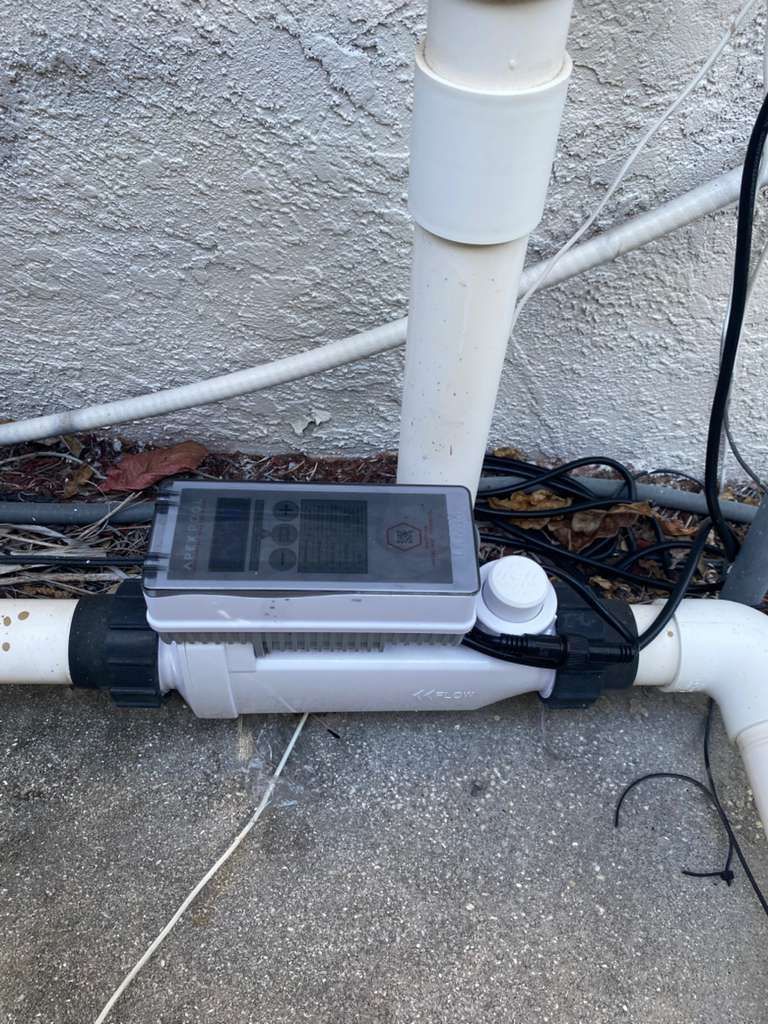 Grey device with a screen and dial on a white pipe, connected to other pipes against a stucco wall.
