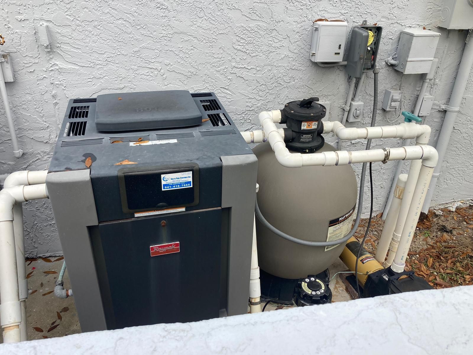 Pool equipment: heater, filter, and pump outside a building. White pipes connect the components.