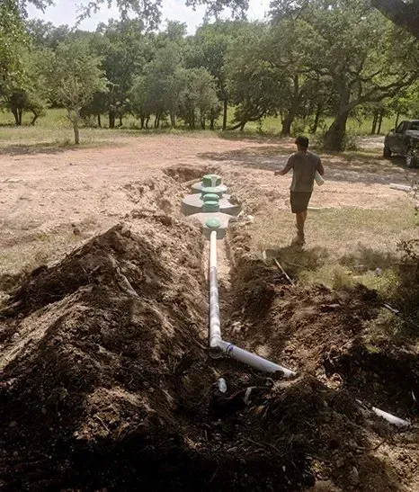 Septic system installation: Man walks toward tanks in a trench, connected by pipes, surrounded by dirt and trees.