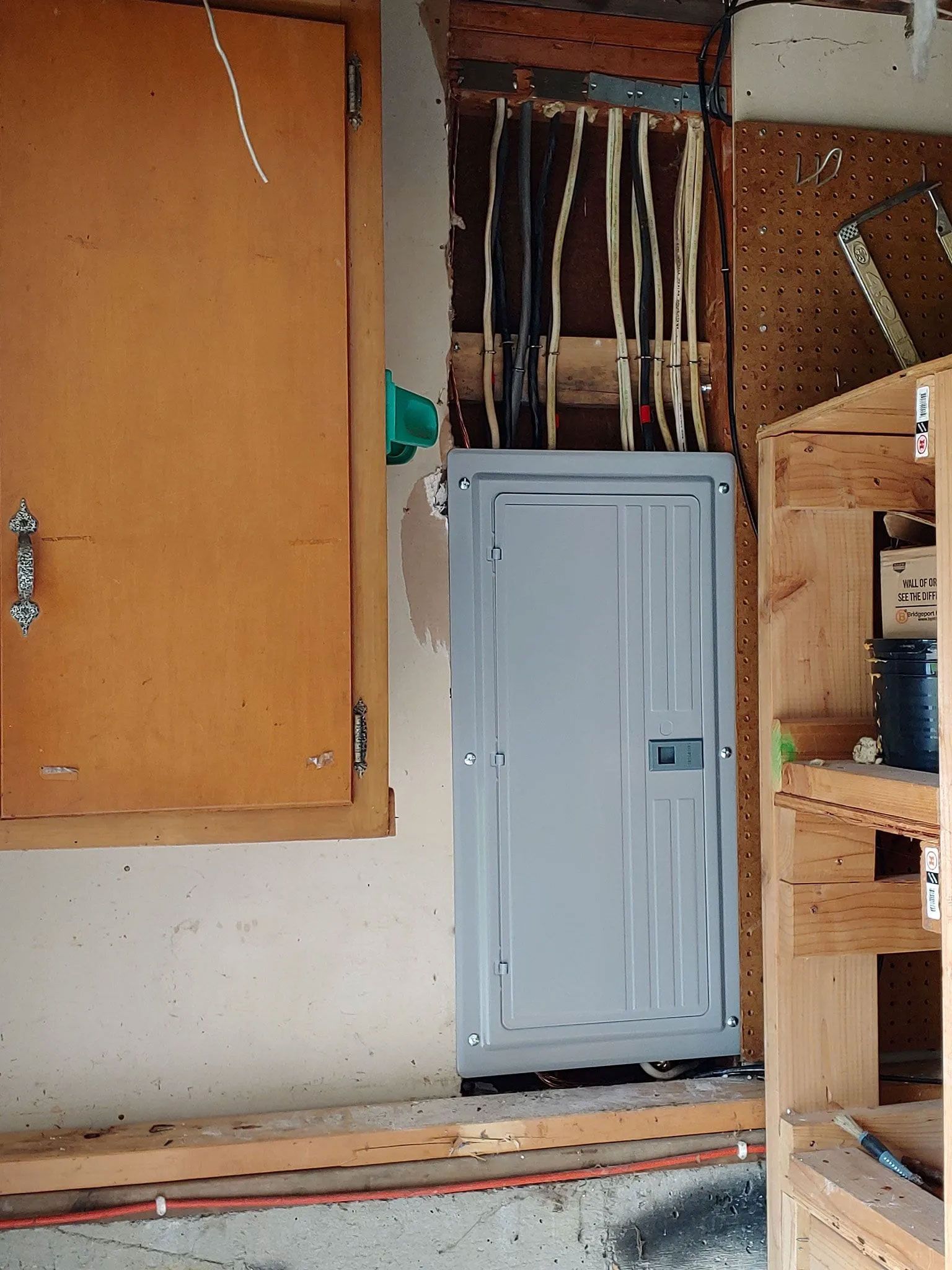 Gray electrical panel in garage, wiring exposed, wooden cabinet and shelving nearby.