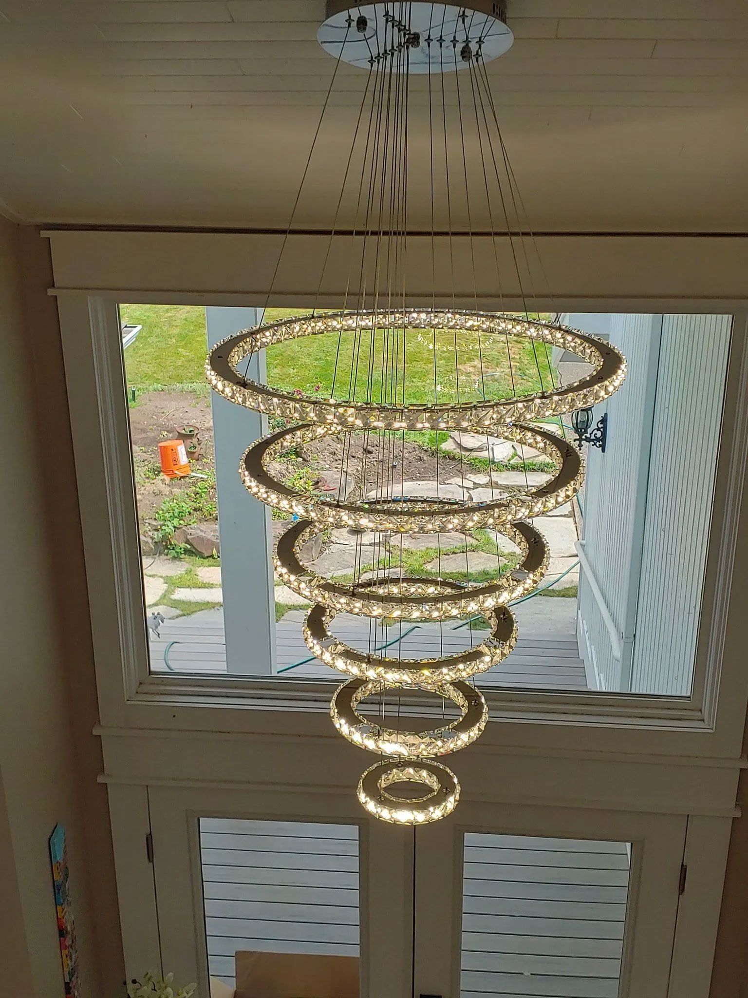Modern chandelier with glowing crystal rings hangs in a bright room, visible through a large window.