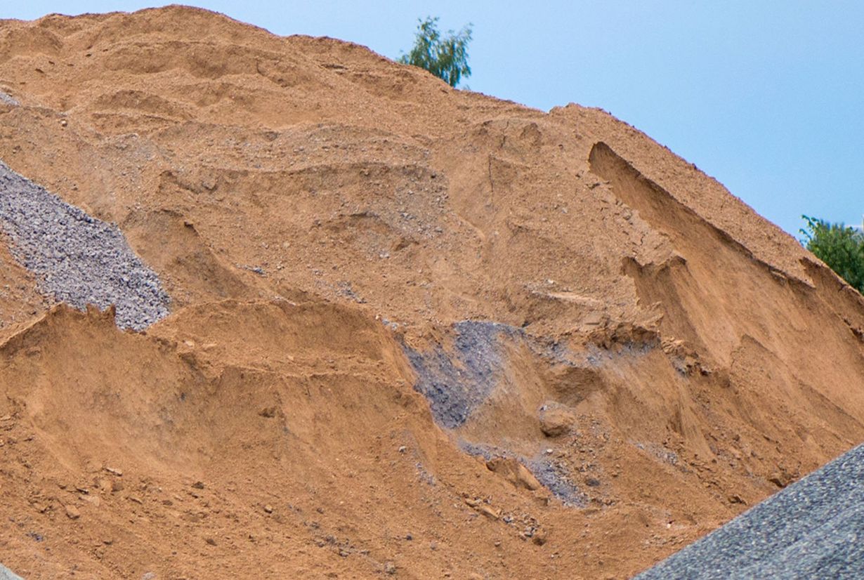 Large pile of reddish-brown sand with some gray gravel visible. Sky and tree in background.