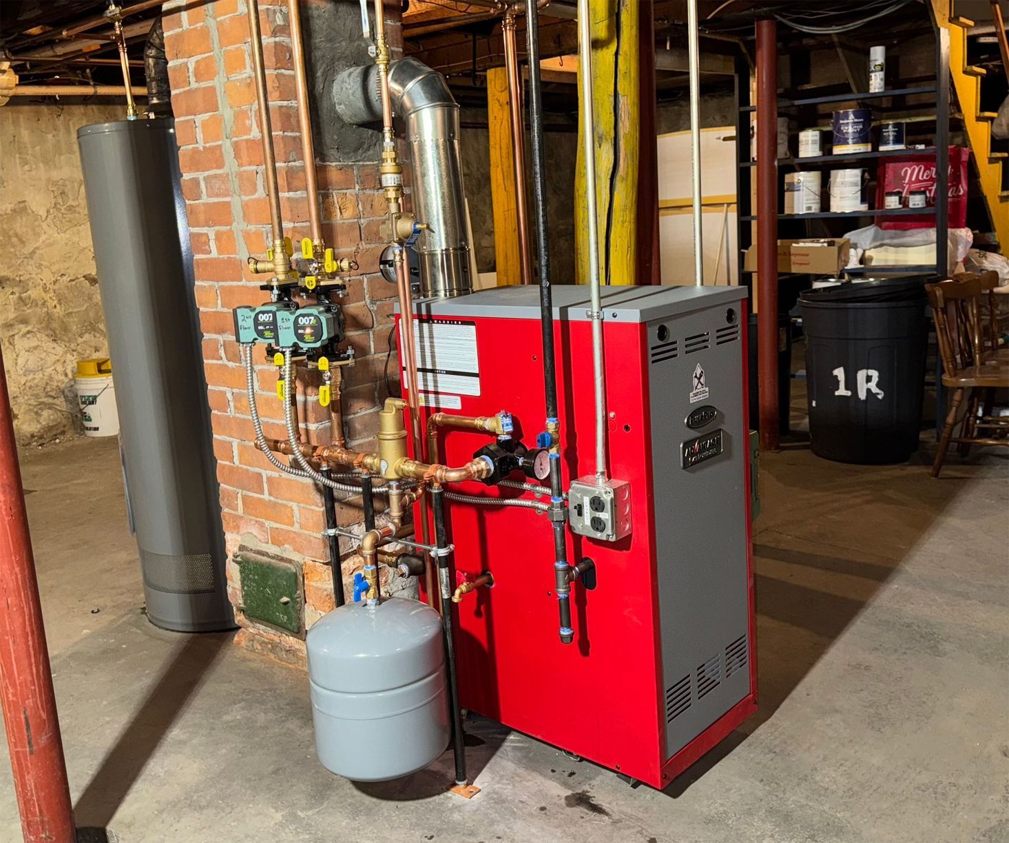A red boiler is sitting in a basement next to a brick wall.