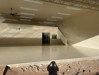 Interior of a large, empty, beige-colored room with a polished floor. Stairs and a doorway are visible. A ceiling fan is mounted.