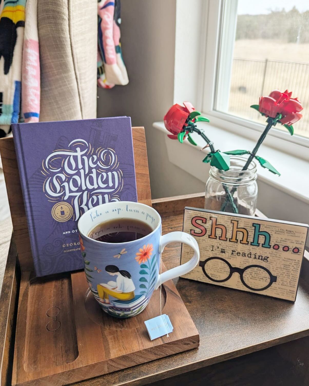 A cup of coffee is sitting on a wooden tray next to a book and a vase of flowers.