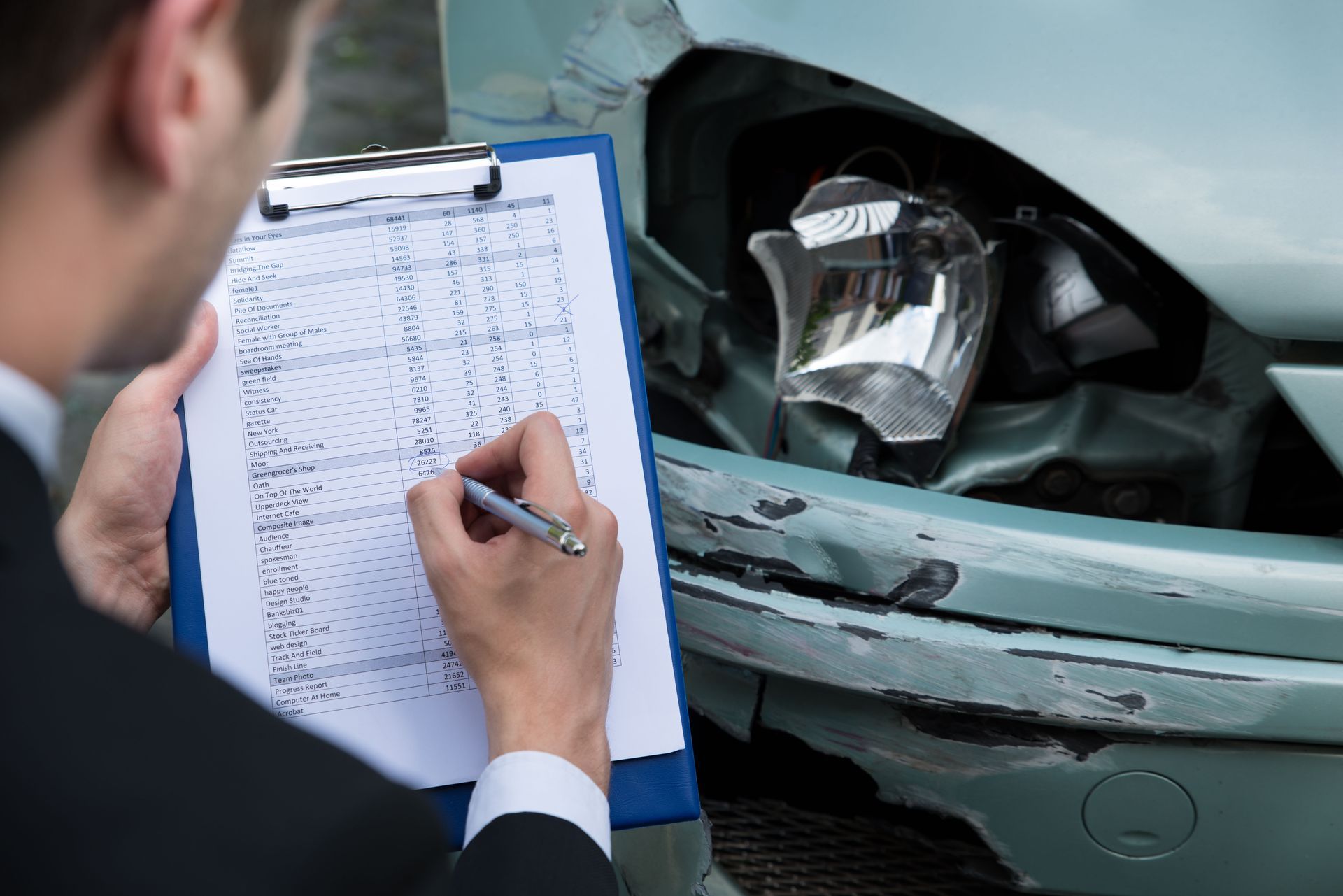 An insurance adjuster assesses car damage. Blue clipboard, pen in hand, looking at a broken headlight and dented car.