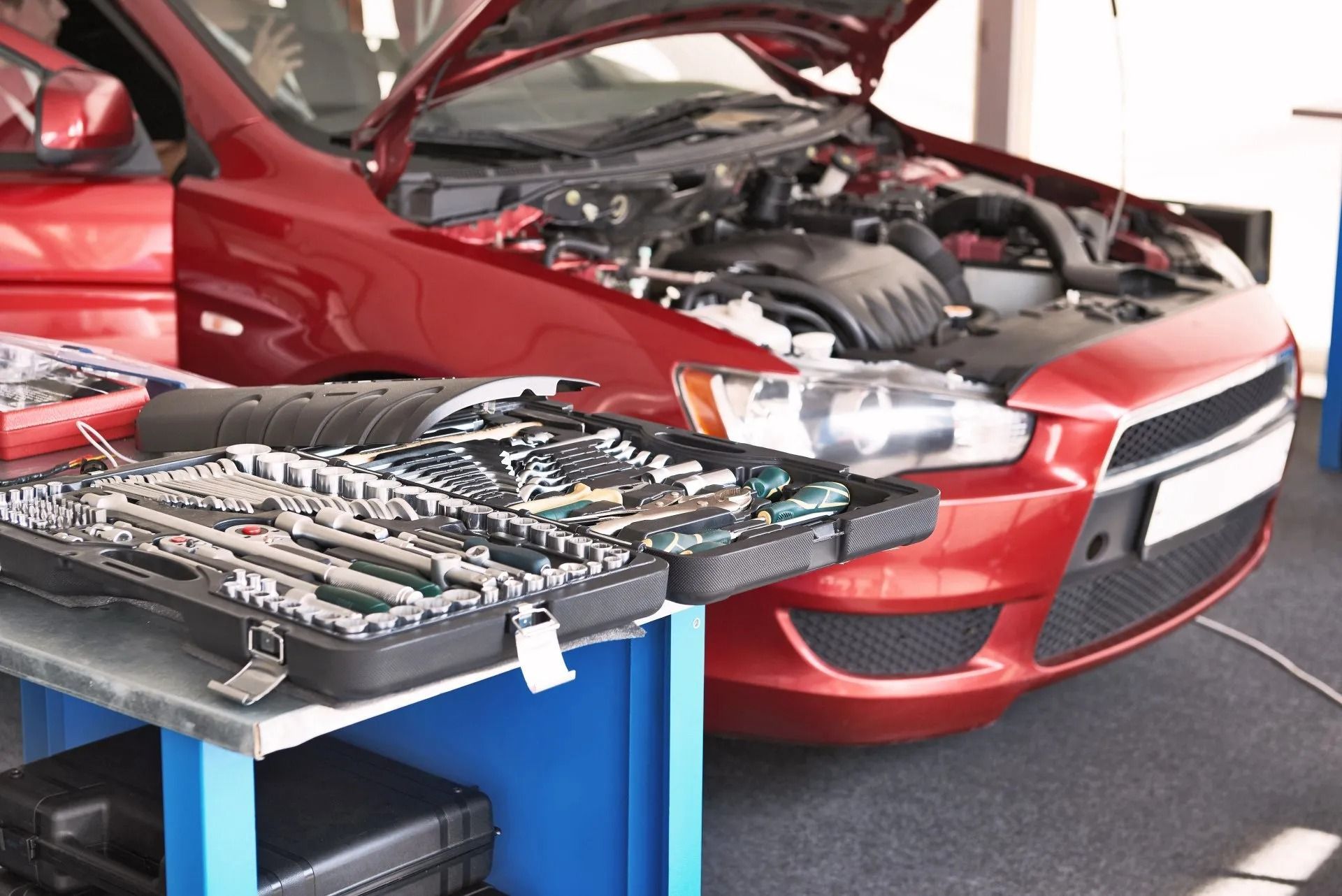 Red car with open hood, tools on blue table in auto repair shop.