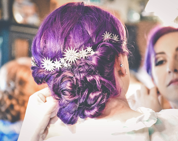 A woman with purple hair is putting on earrings in front of a mirror.
