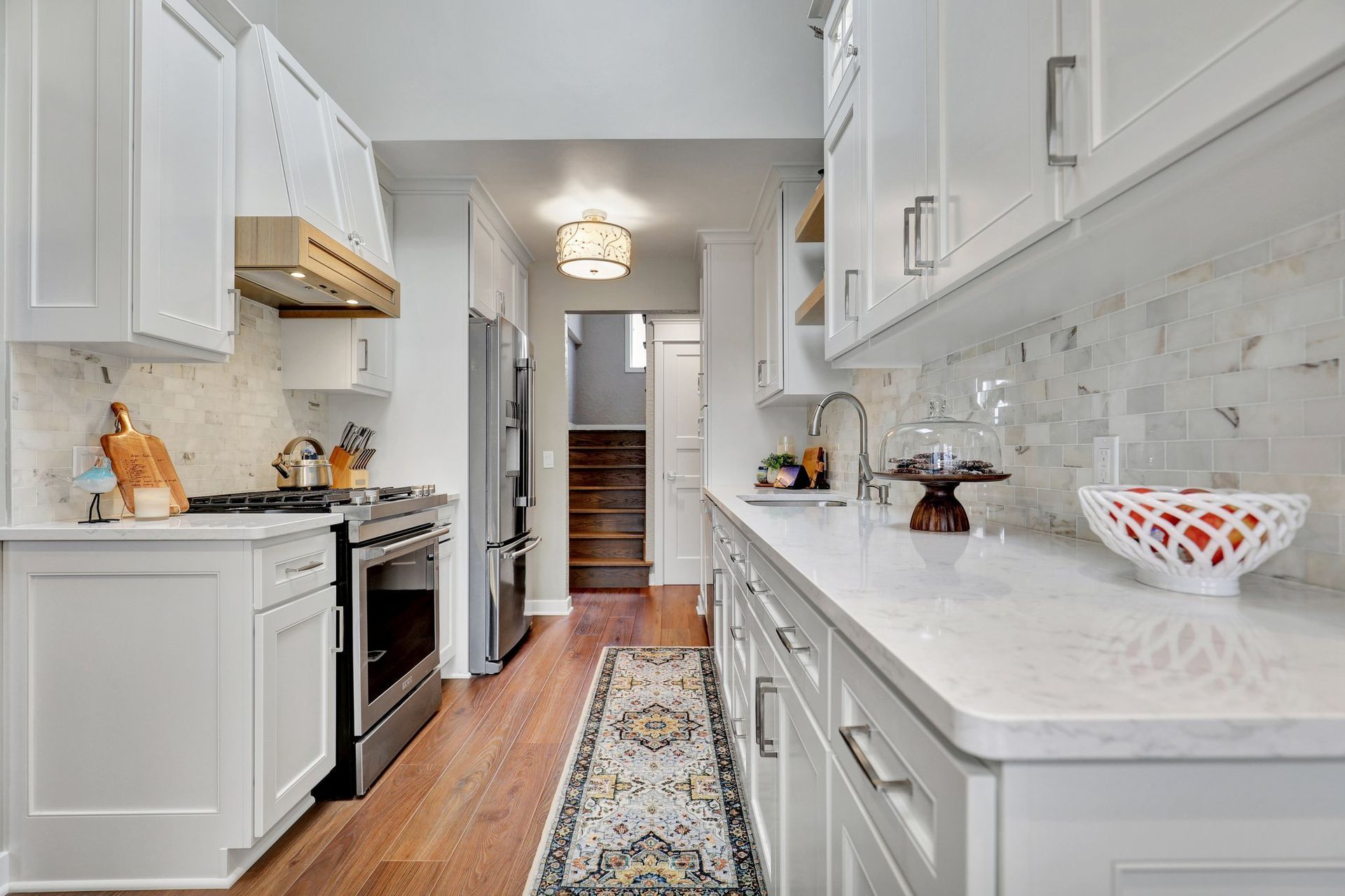 Bright white kitchen with marble counters, stainless stove, and patterned runner leading to a hallway.