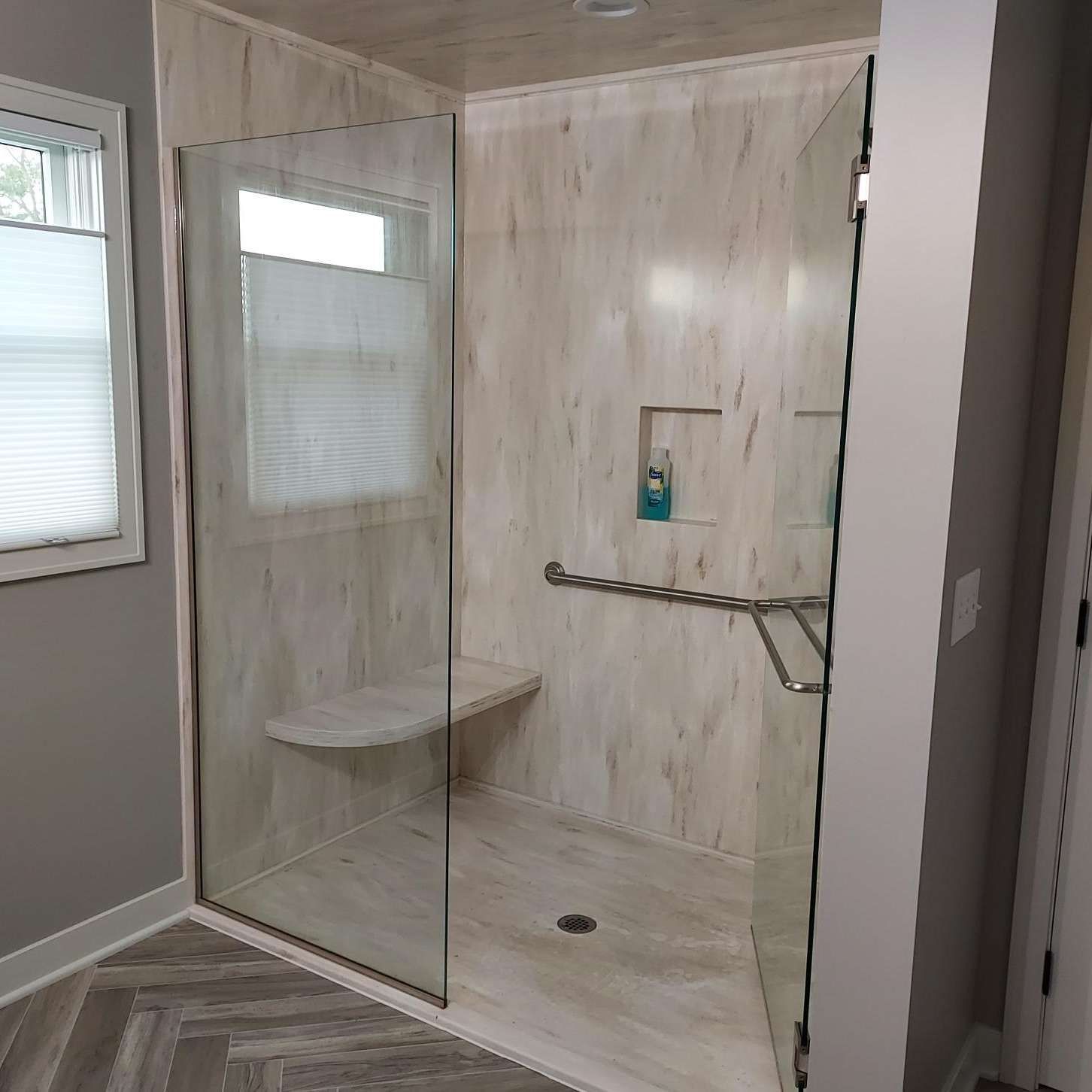 Modern bathroom with glass shower enclosure, bench seat, grab bars, and light wood-look tile walls.