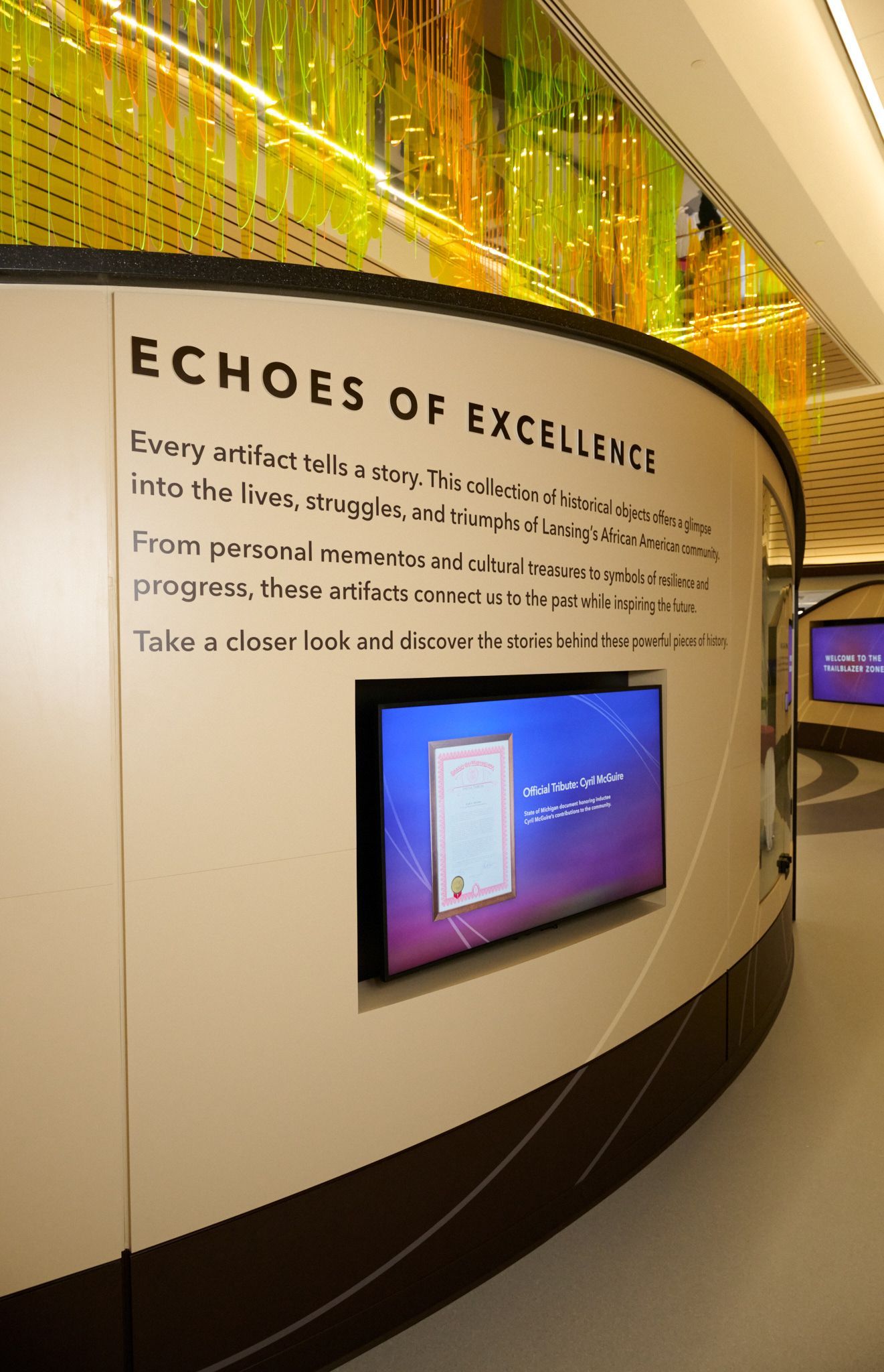 Curved museum wall display titled “Echoes of Excellence” with text panel and embedded screen