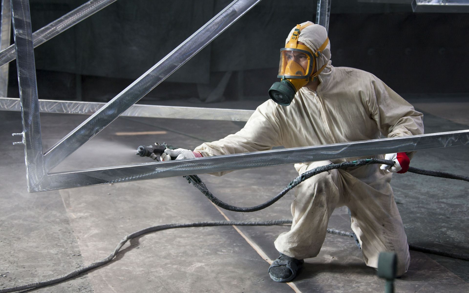 Person in protective gear sprays a metal frame in an industrial setting.