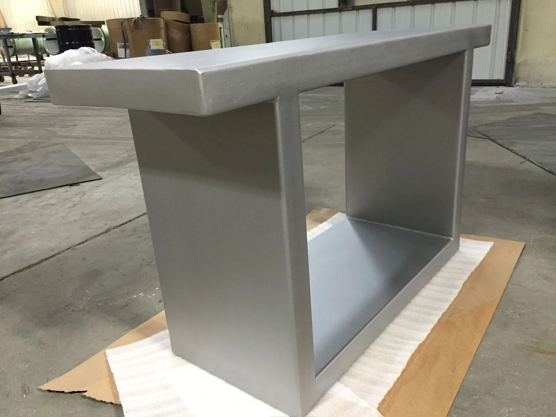 A metal, rectangular table with a hollow center sits on a cardboard sheet indoors.
