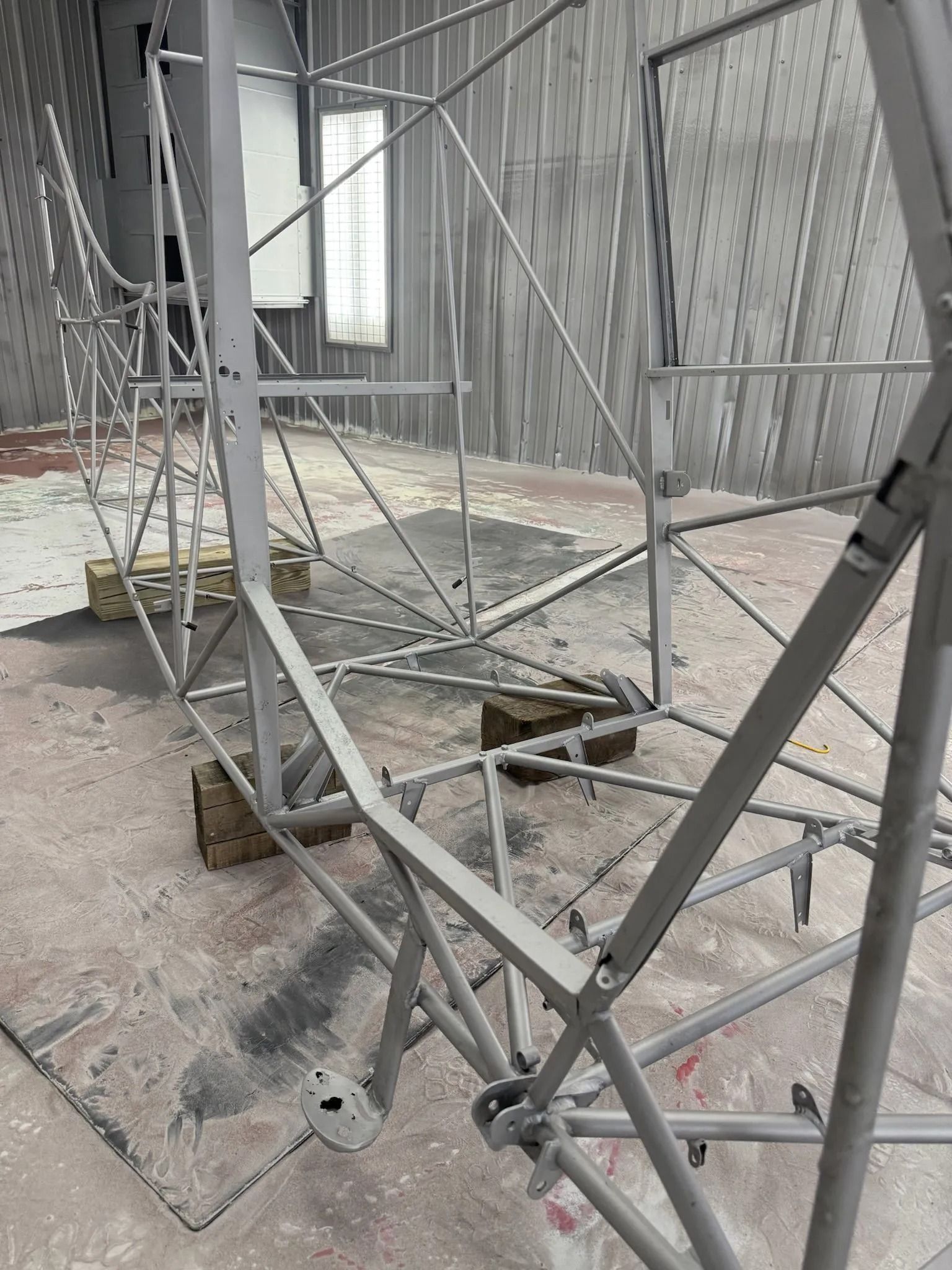 Gray painted aircraft fuselage frame on wooden blocks in a spray booth.