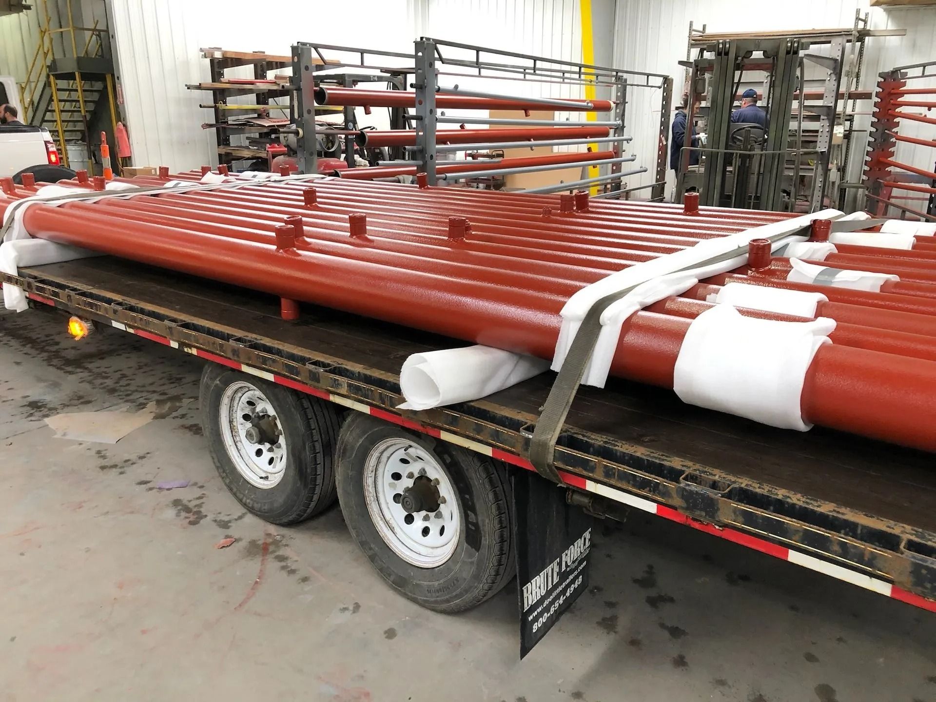 Red pipes secured with straps on a flatbed trailer, inside a warehouse.