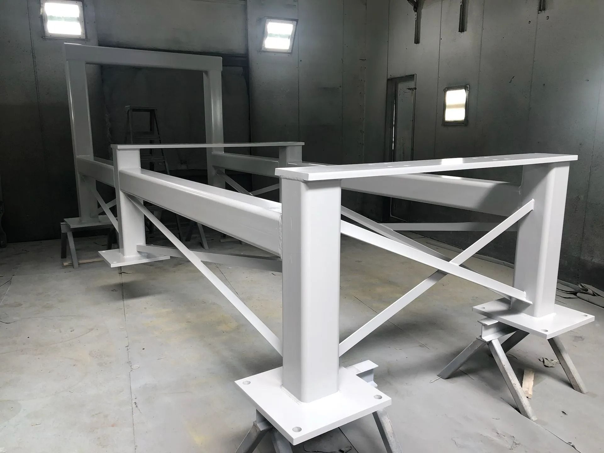 White-painted metal structure in a spray booth. Square support posts with cross bracing and horizontal beams.