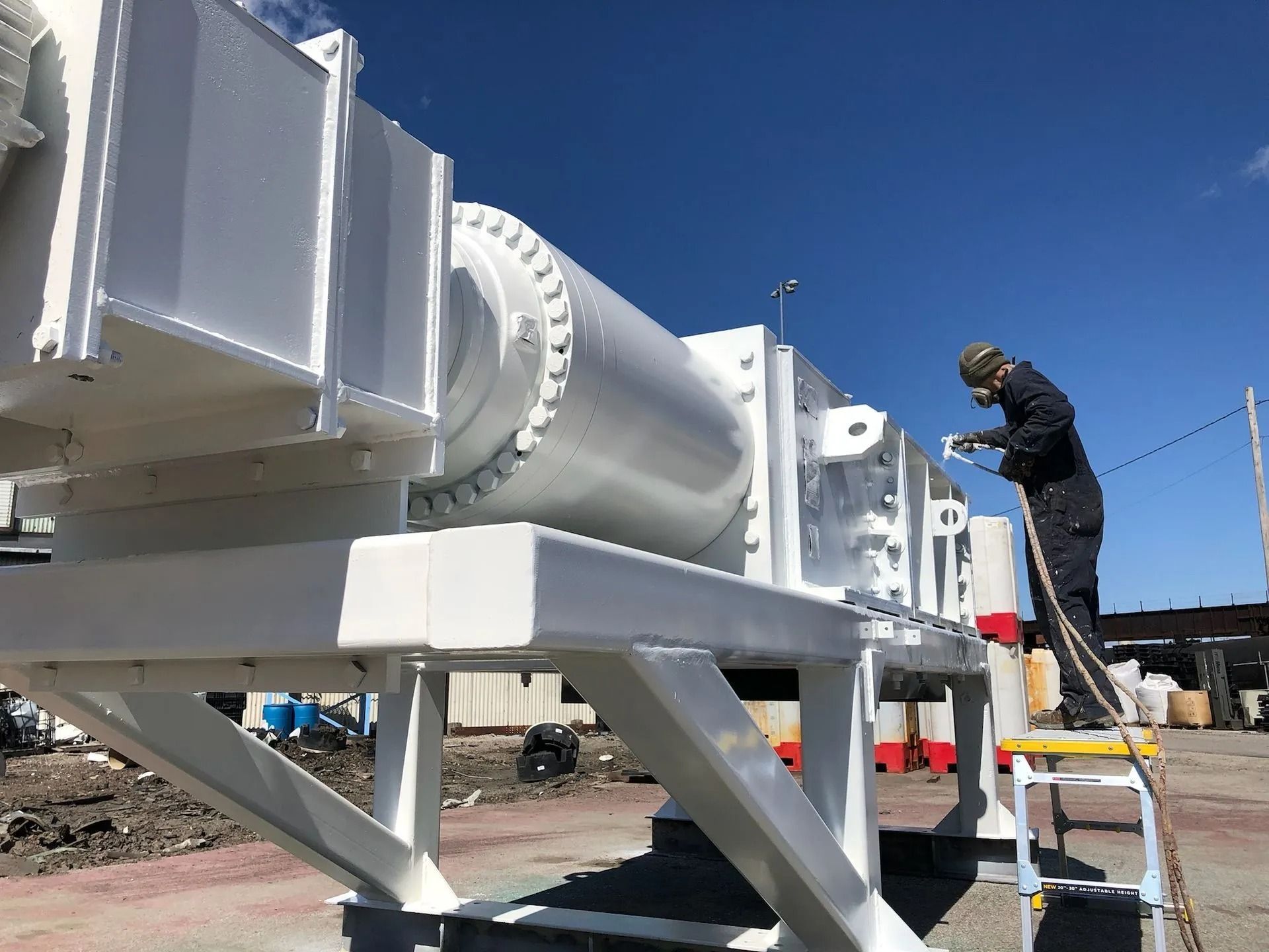 Person on a step ladder painting a large white industrial machine outdoors. Blue sky background.