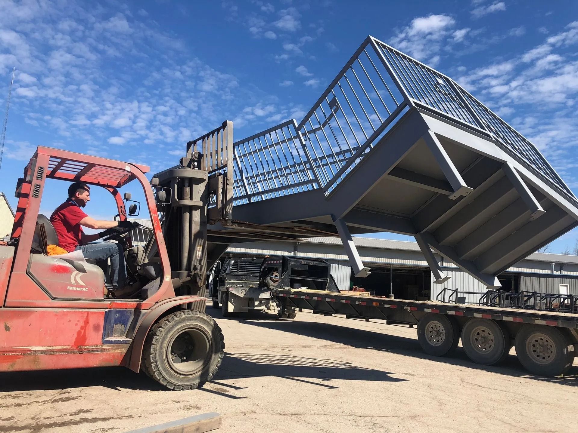 Forklift lifting a large metal structure onto a flatbed trailer under a blue sky. A person steers the forklift.