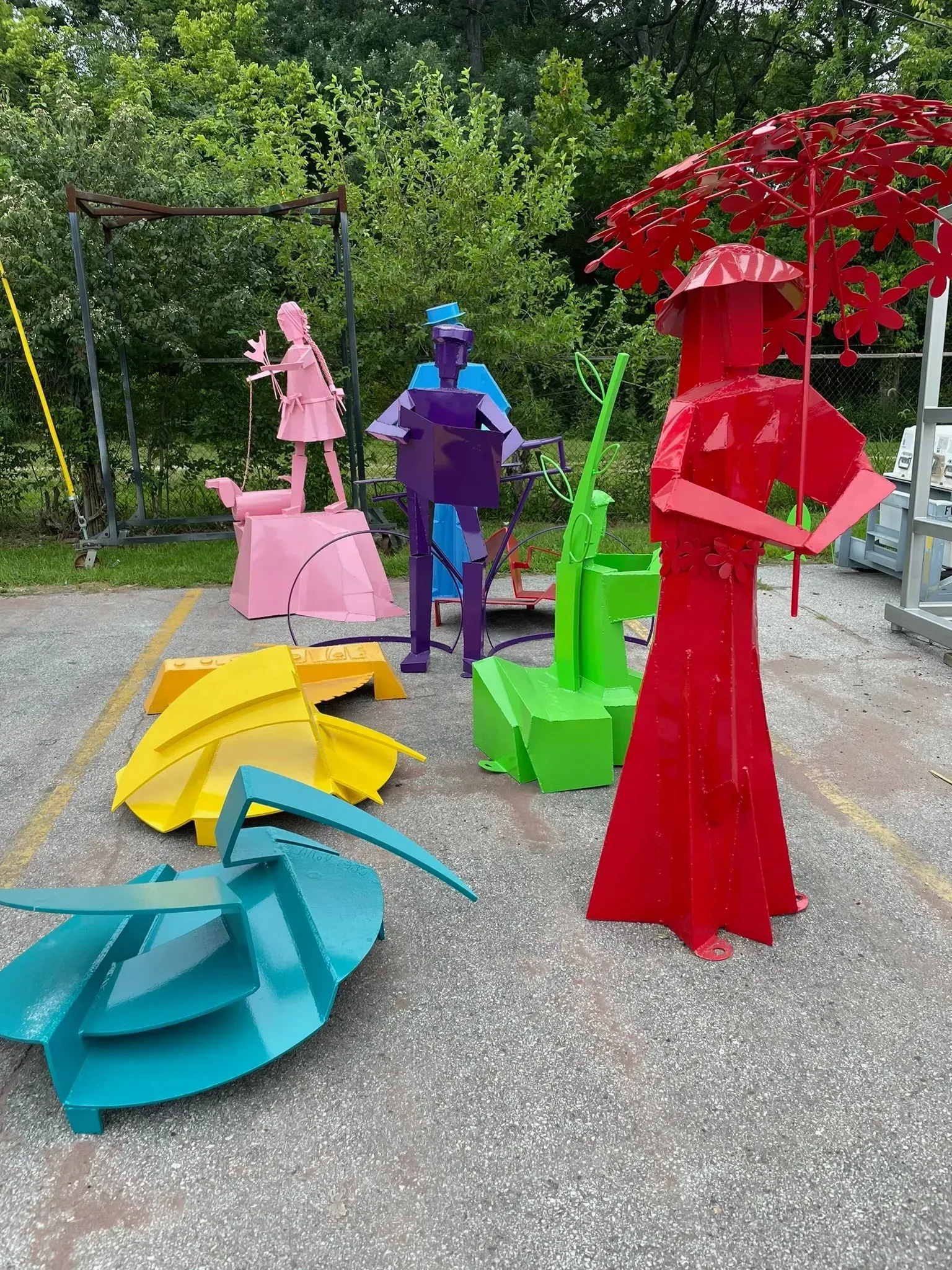 Colorful metal sculptures in a parking lot, including figures and abstract shapes in shades of red, purple, green, blue, yellow, and pink.