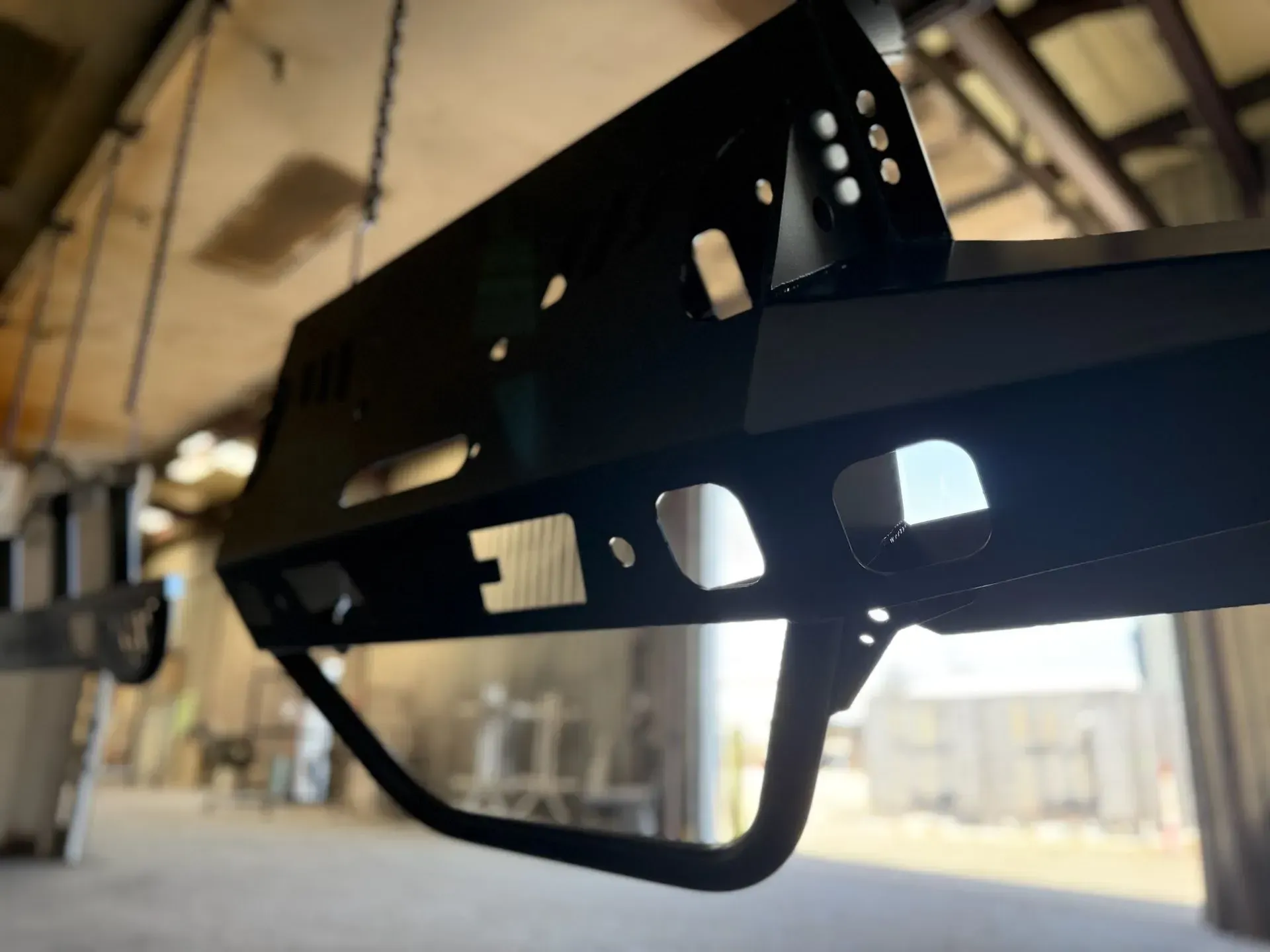 Black metal bumper with rectangular and arched openings, hanging in a workshop.