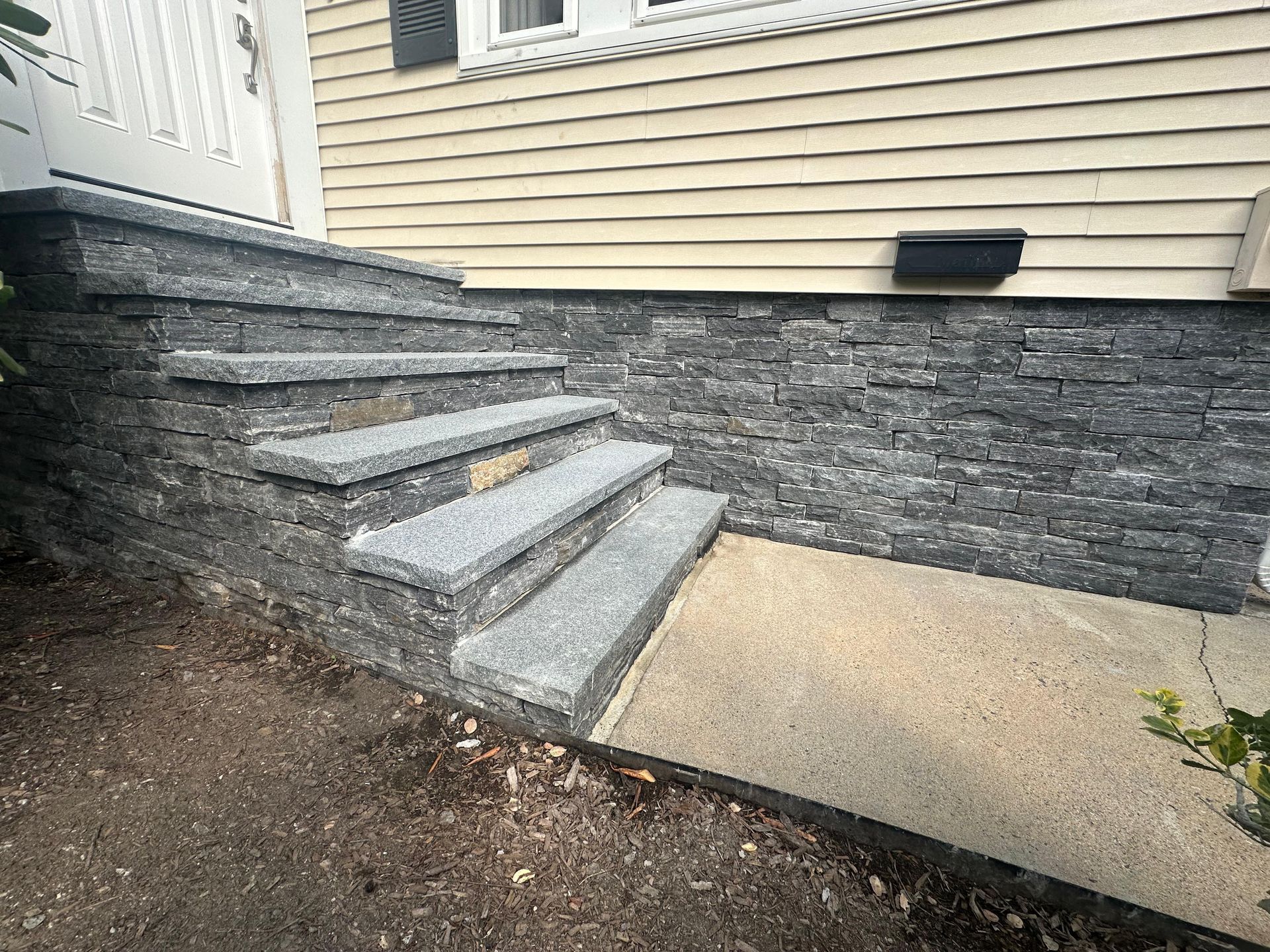 Concrete steps leading to a house entrance beside a stone retaining wall and beige siding