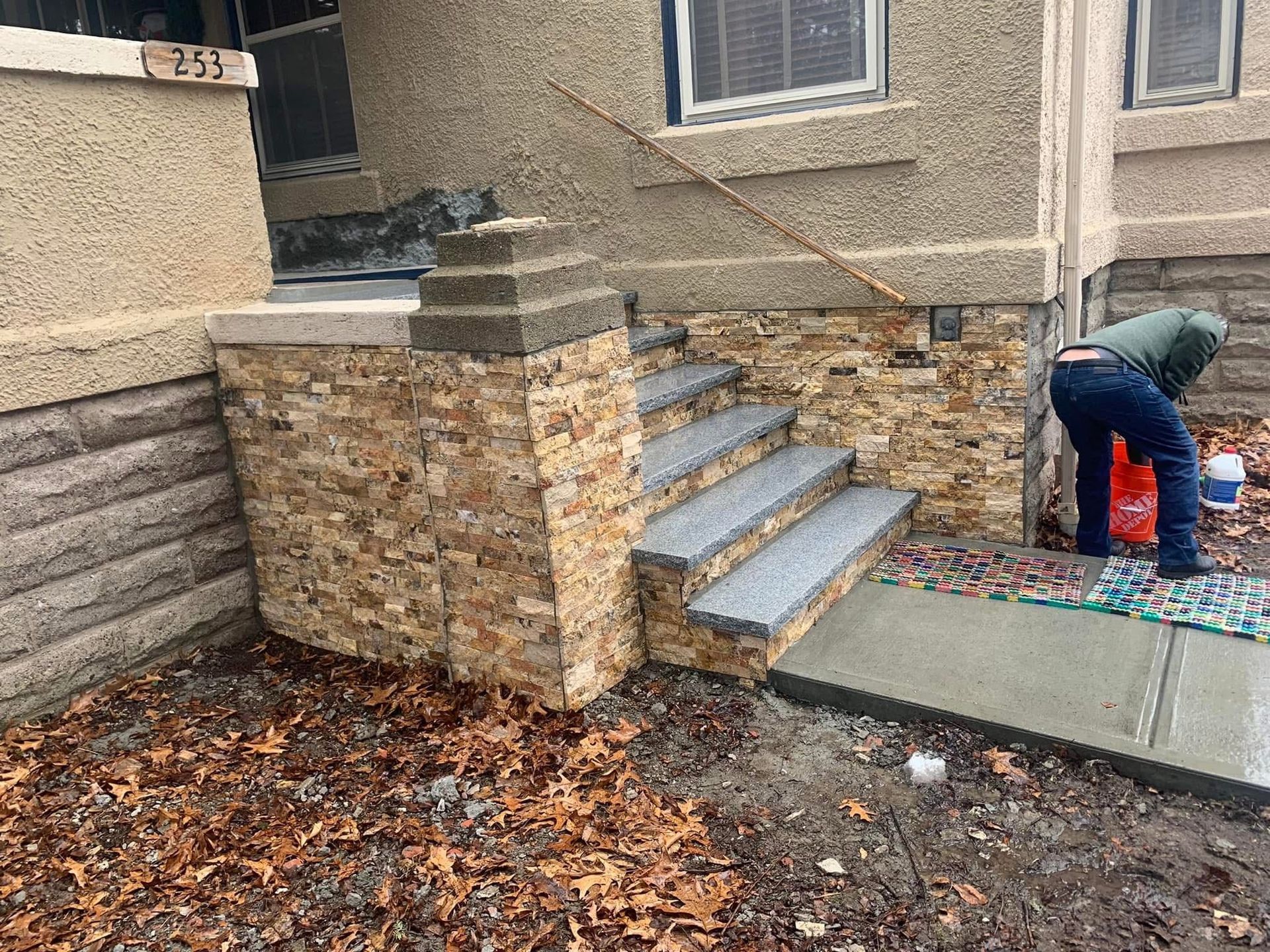 Worker cleaning a front stoop and walkway beside a stone-and-siding house in wet, leaf-covered weather
