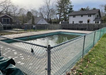 Chain-link fenced backyard swimming pool with covered water, surrounded by houses and bare trees