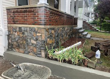 Brick and stone house porch with steps, small garden bed, and sidewalk planter in front of a residential yard