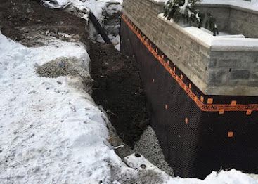 Snow-covered yard beside a damaged brick retaining wall with a deep erosion trench.