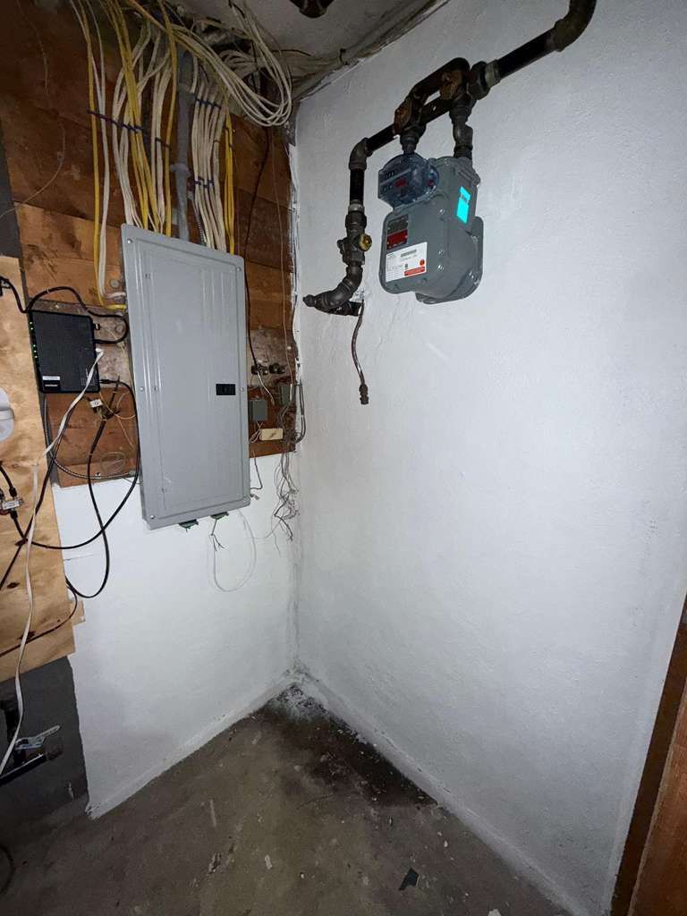 Small utility room with white walls, exposed wiring, electrical panels, and concrete floor.