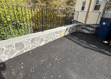 Stone retaining wall beside black asphalt driveway next to a house and blue bin, with black metal fence above