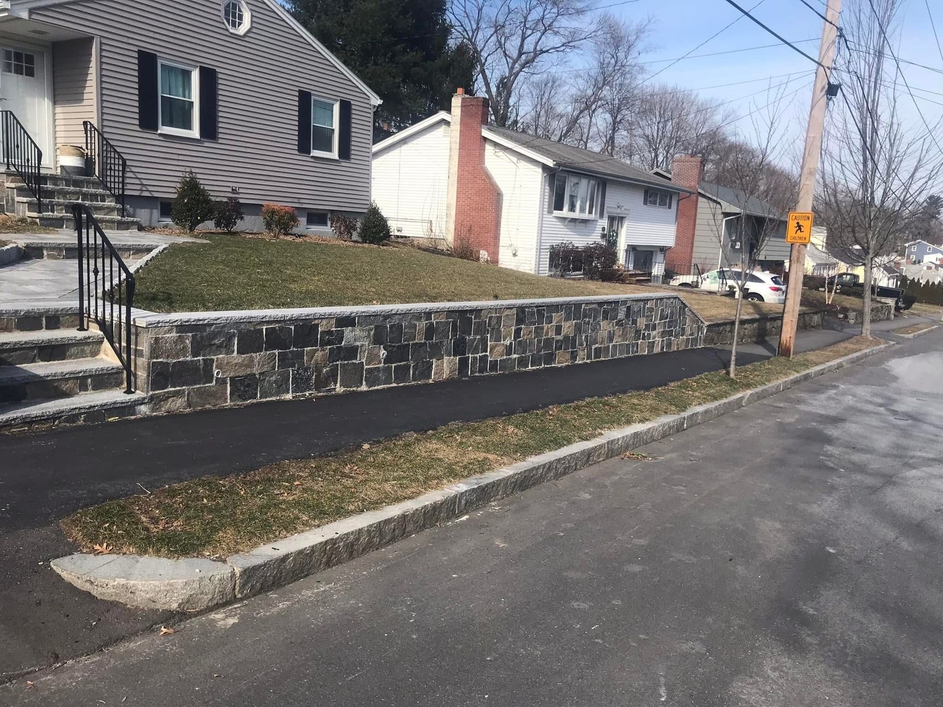 Residential street with stone retaining wall, houses, and a sidewalk beside the road