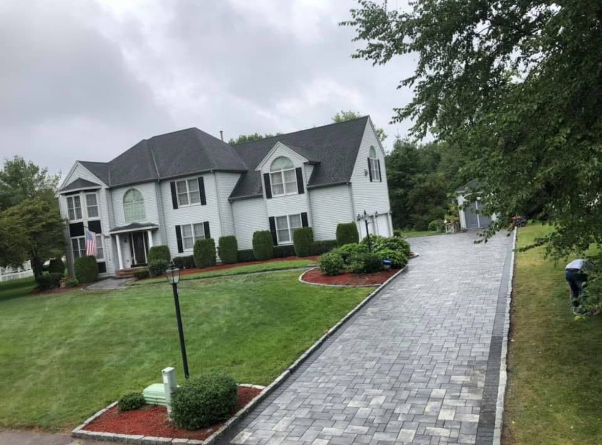 Large suburban house with a long stone driveway, green lawn, and landscaped front yard under overcast skies