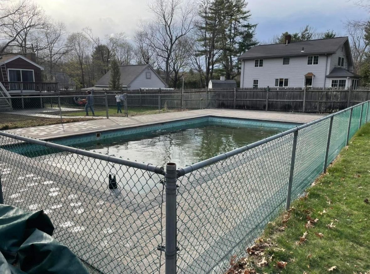 Empty fenced backyard swimming pool beside houses and trees on an overcast day